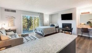 A modern hotel room with a large bed, a dining area, a fireplace, and a balcony with a view of trees.