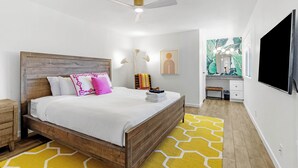 Une chambre avec un lit en bois, du linge de lit blanc, un tapis à motifs jaunes, un ventilateur de plafond, une table de chevet et un téléviseur fixé au mur.