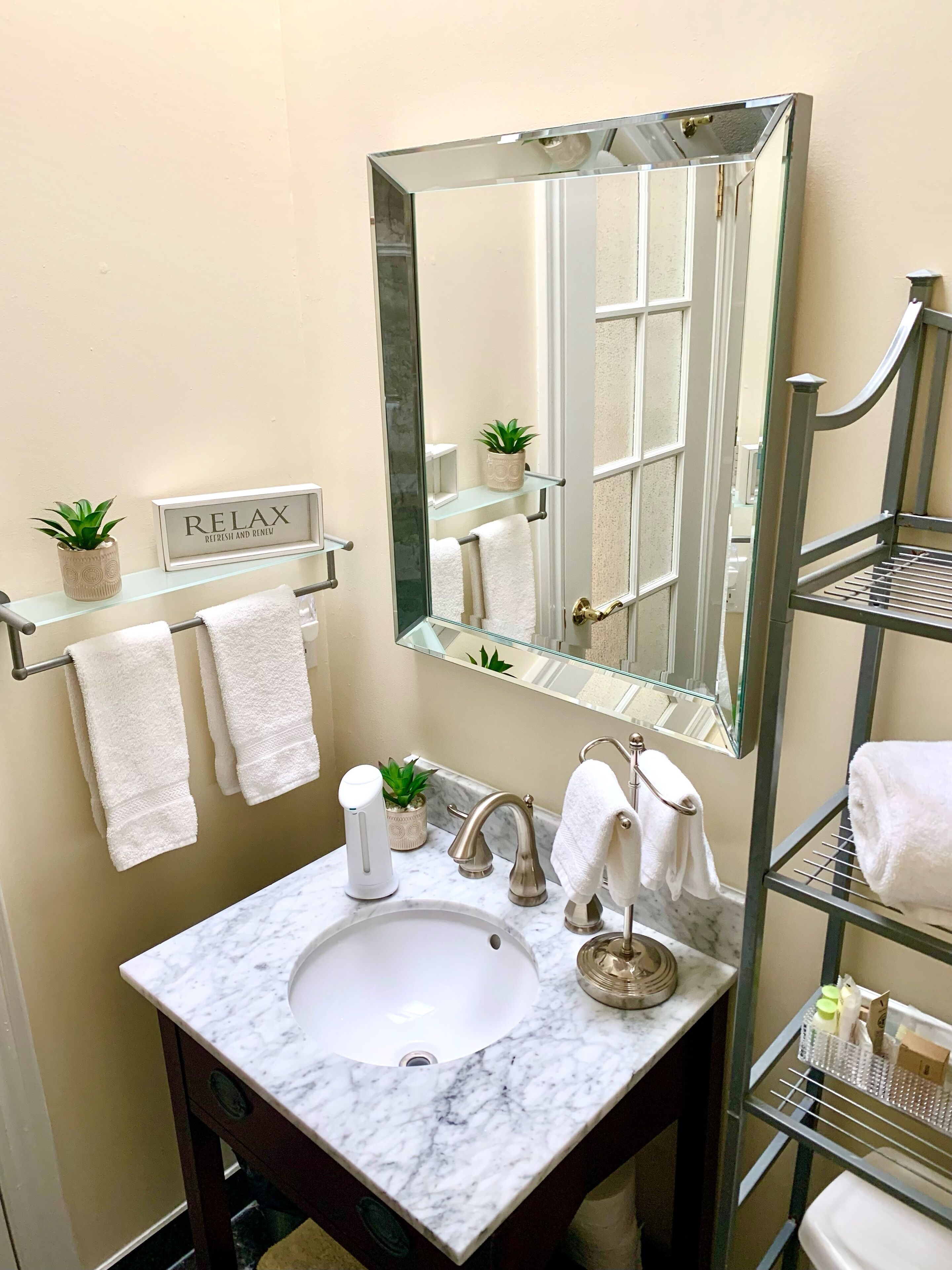 A bathroom with a marble sink, a large mirror, a towel rack with two towels, a shelf with a plant, and a sign that says "RELAX".