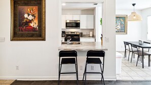 Une cuisine moderne avec des armoires blanches, un four à micro-ondes en acier inoxydable et un comptoir en marbre. On y trouve deux tabourets de bar noirs et une table à manger avec des chaises dans l’espace repas adjacent.