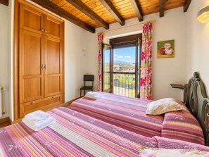 Un dormitorio con un amplio armario de madera, una cama con ropa de cama a rayas, una silla y vistas a través de una ventana con cortinas.