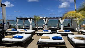 Um espaço de relaxamento à beira-mar com várias espreguiçadeiras, cada uma com colchão e almofadas, sob toldos brancos.