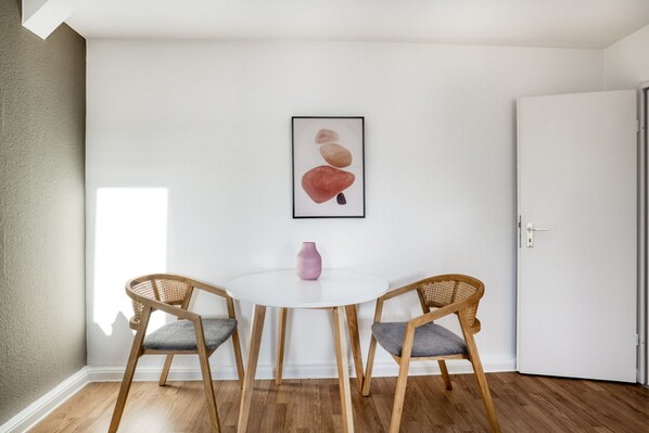 Un comedor con una mesa redonda, dos sillas de mimbre, un florero rosa y un cuadro abstracto enmarcado en la pared.