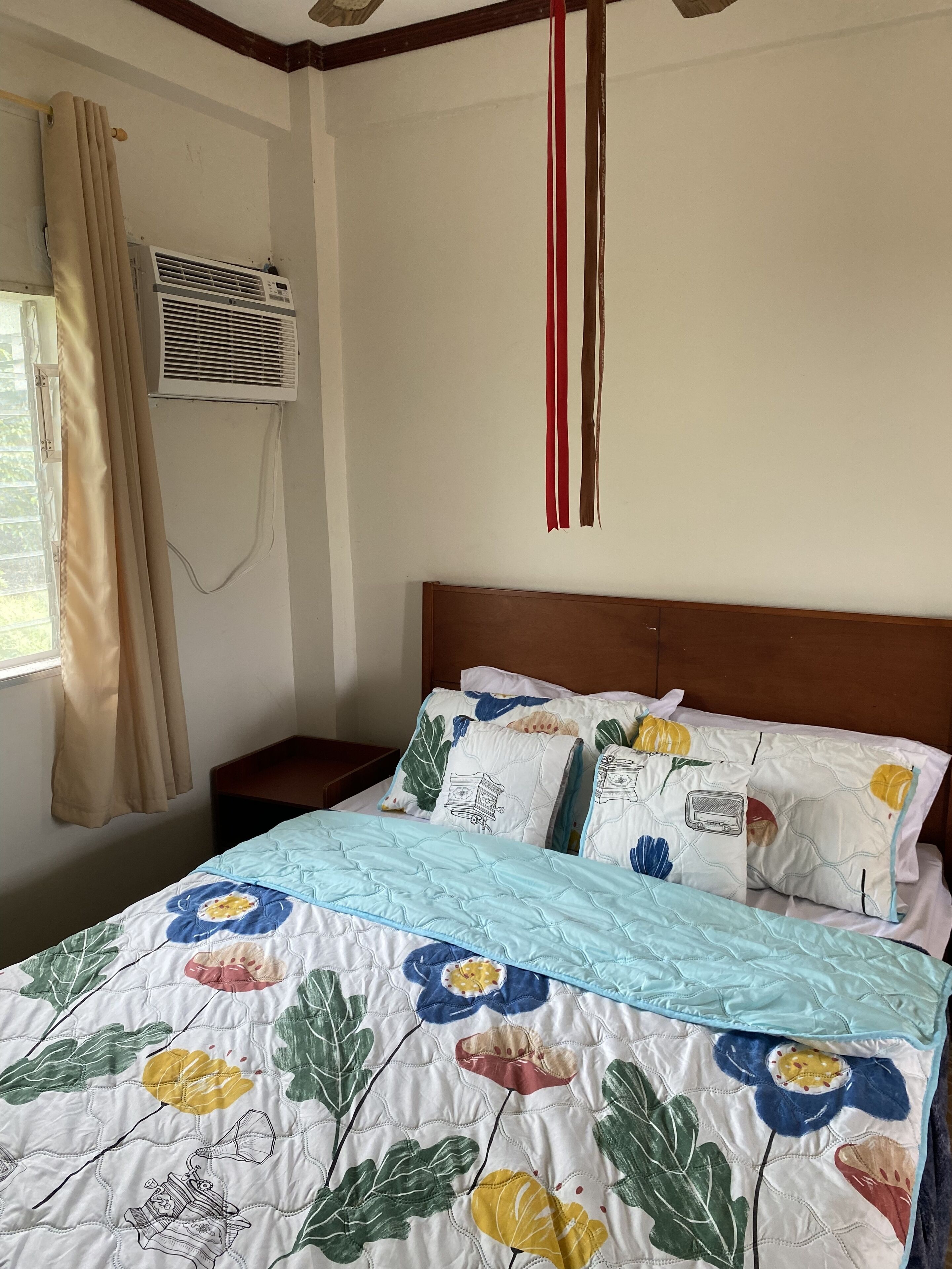A bedroom with a bed, a window with curtains, an air conditioning unit, and a wooden headboard.