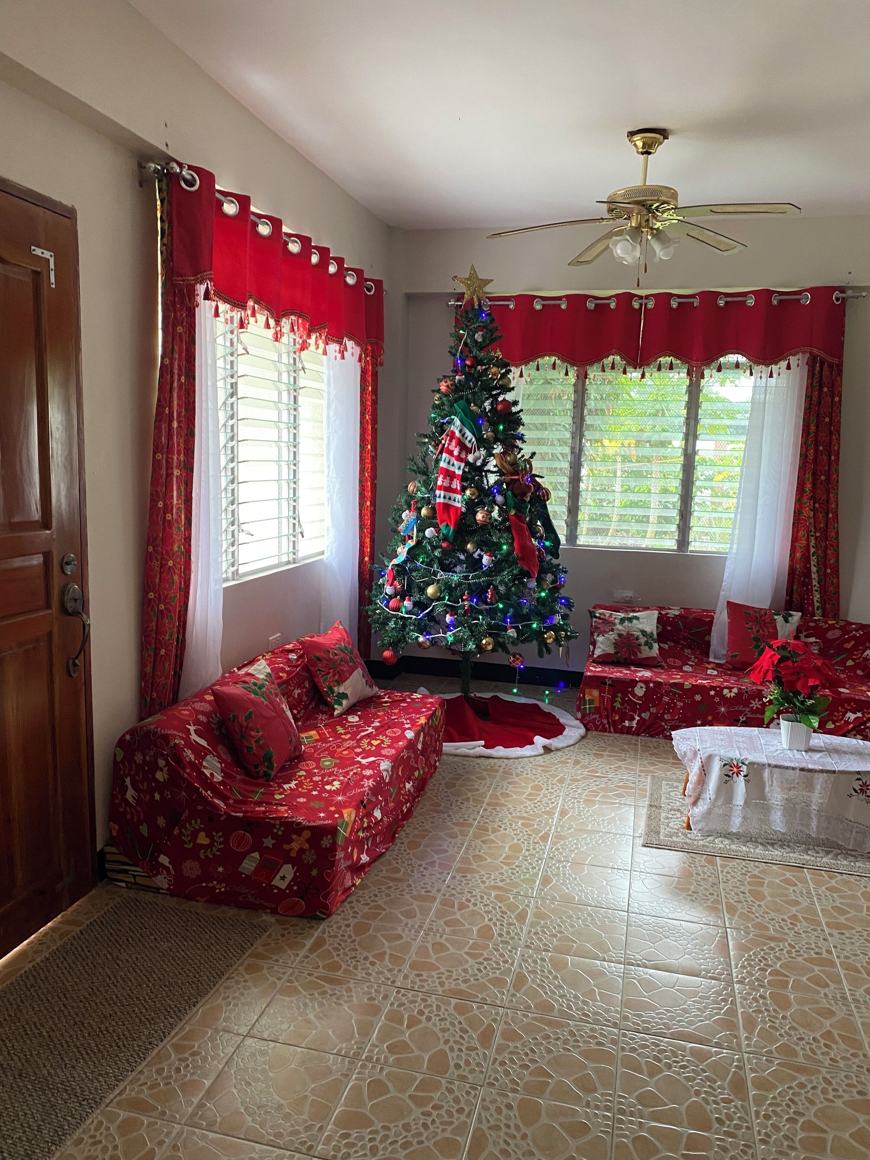 Ein Wohnzimmer mit Weihnachtsbaum, roten Vorhängen, einer Holztür und einem Fliesenboden.