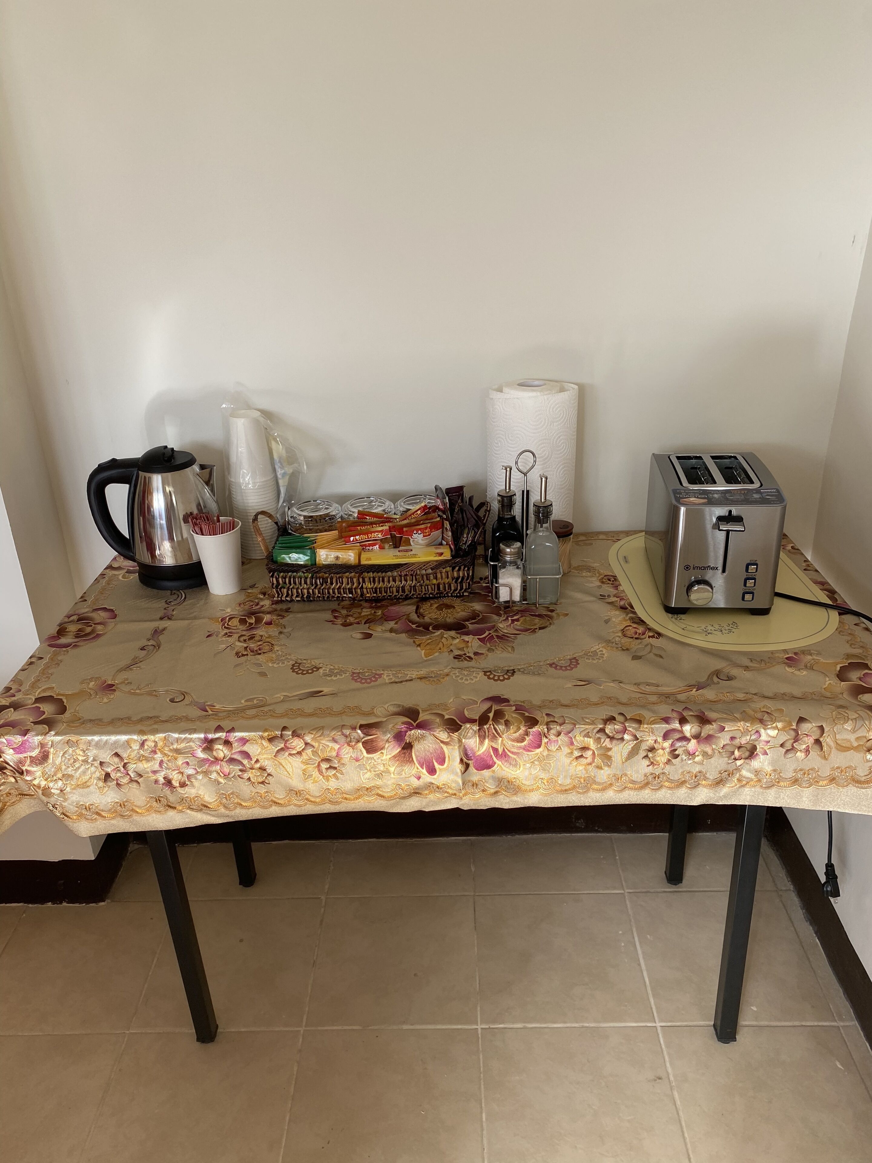 Una mesa con mantel floral, una tetera, un tostador, una cesta de aperitivos y un rollo de toallas de papel.
