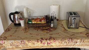 Una mesa con mantel floral, una tetera, un tostador, una cesta de aperitivos y un rollo de toallas de papel.