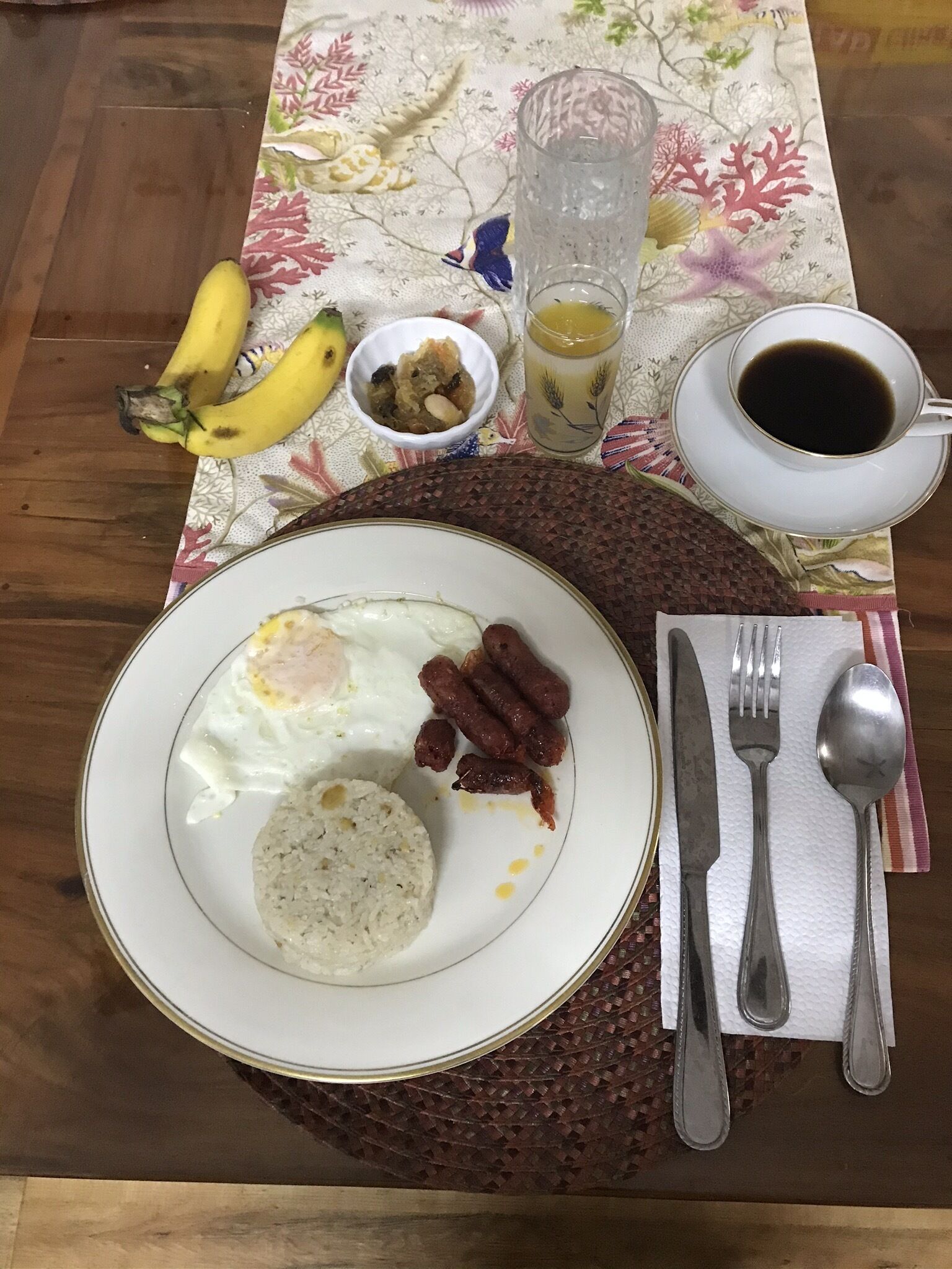 A breakfast setting with a plate of eggs, sausages, and a side dish, accompanied by bananas, a glass of juice, and a cup of coffee on a table with a floral tablecloth.