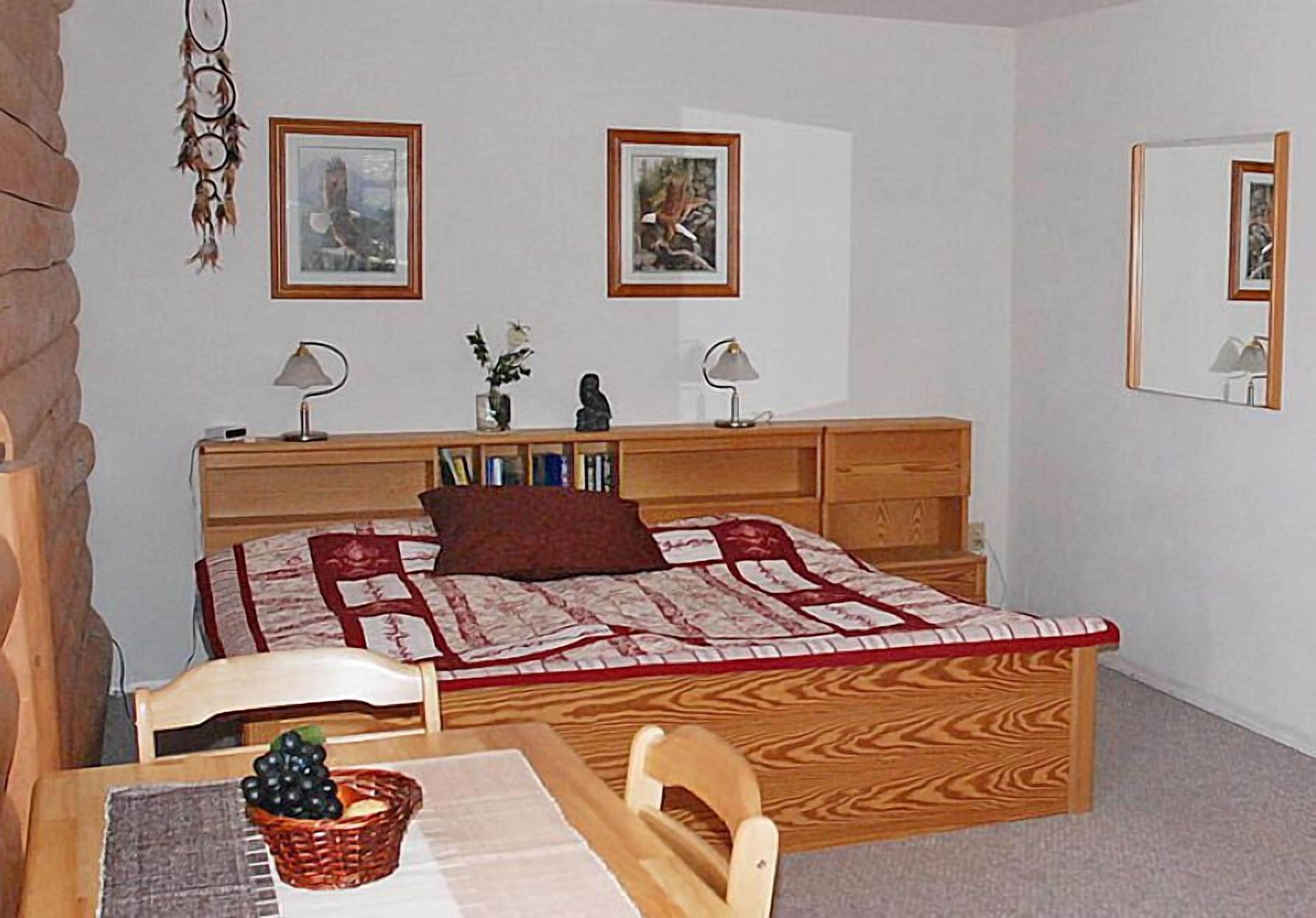 Une chambre à coucher avec un lit, des tables de chevet, un bureau et une chaise. Il y a deux tableaux encadrés au mur, un miroir et un attrape-rêves.