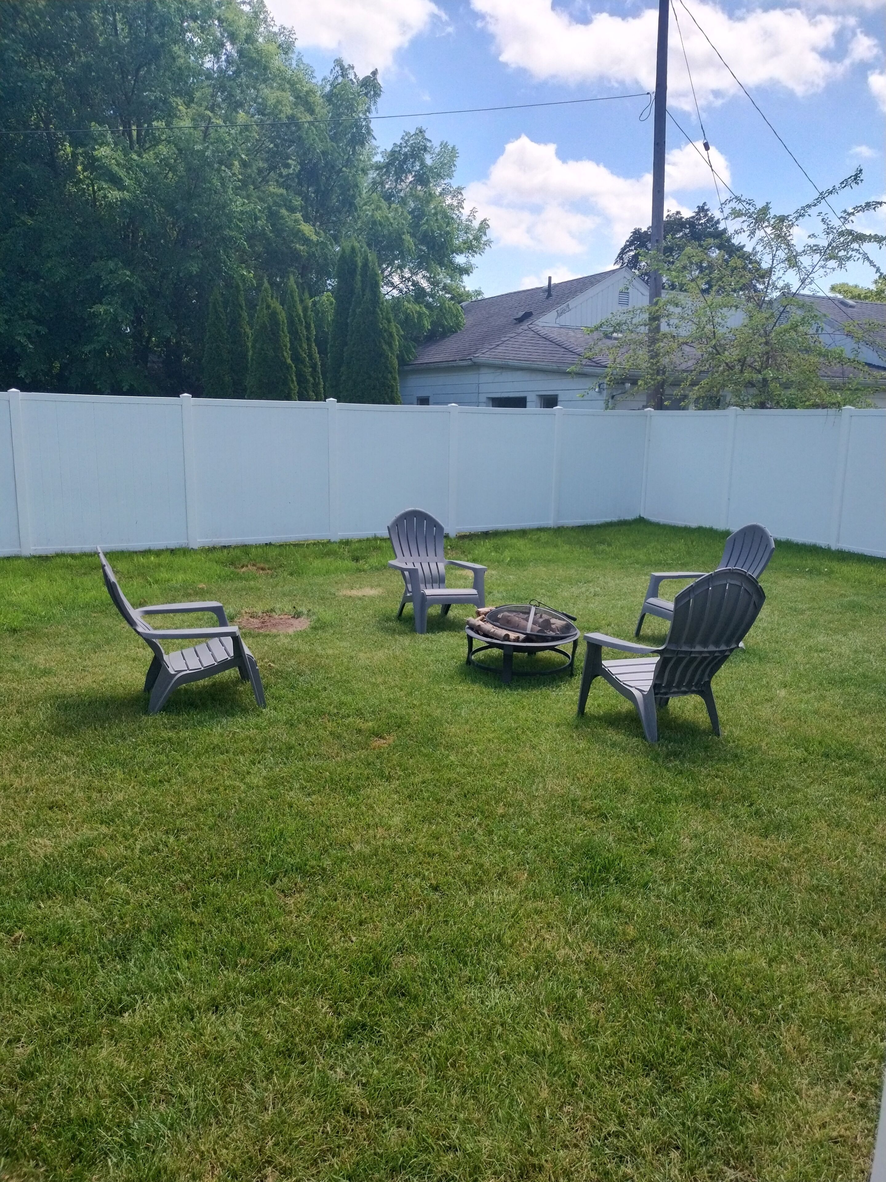 A backyard with a white fence, three Adirondack chairs, and a fire pit.