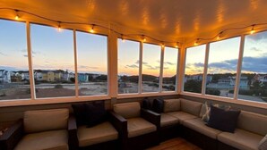 Ein überdachter Patio-Bereich mit gepolsterten Sitzgelegenheiten und großen Fenstern, die einen Blick auf eine Wohngegend bieten.