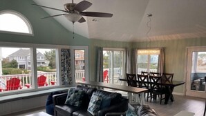 Ein Wohnzimmer mit einer schwarzen Ledersofa, einem Esstisch mit Stühlen, einem Deckenventilator und großen Fenstern mit Blick auf die Nachbarschaft.
