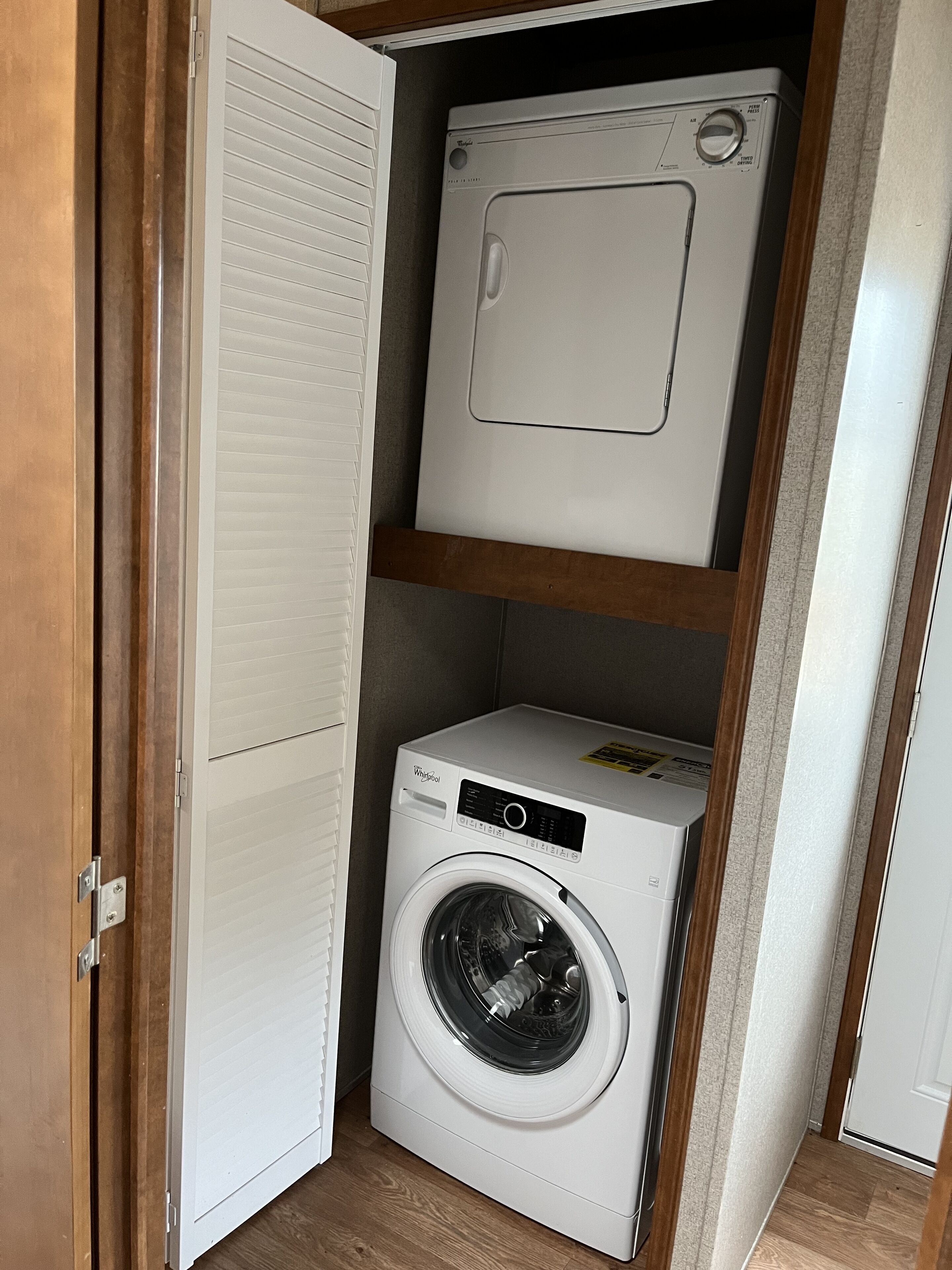 A compact laundry room with a front-loading washer and dryer, wooden shelving, and a door with a louvered design.