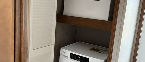 A compact laundry room with a front-loading washer and dryer, wooden shelving, and a door with a louvered design.