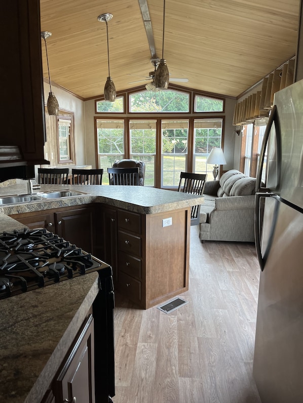 A kitchen with a central island, wooden cabinets, a gas stove, and a refrigerator. There are large windows providing natural light and a view of greenery outside.