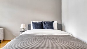 Una cama bien hecha con una colcha gris y blanca a cuadros, dos almohadas y una lámpara blanca sobre una mesita de noche.