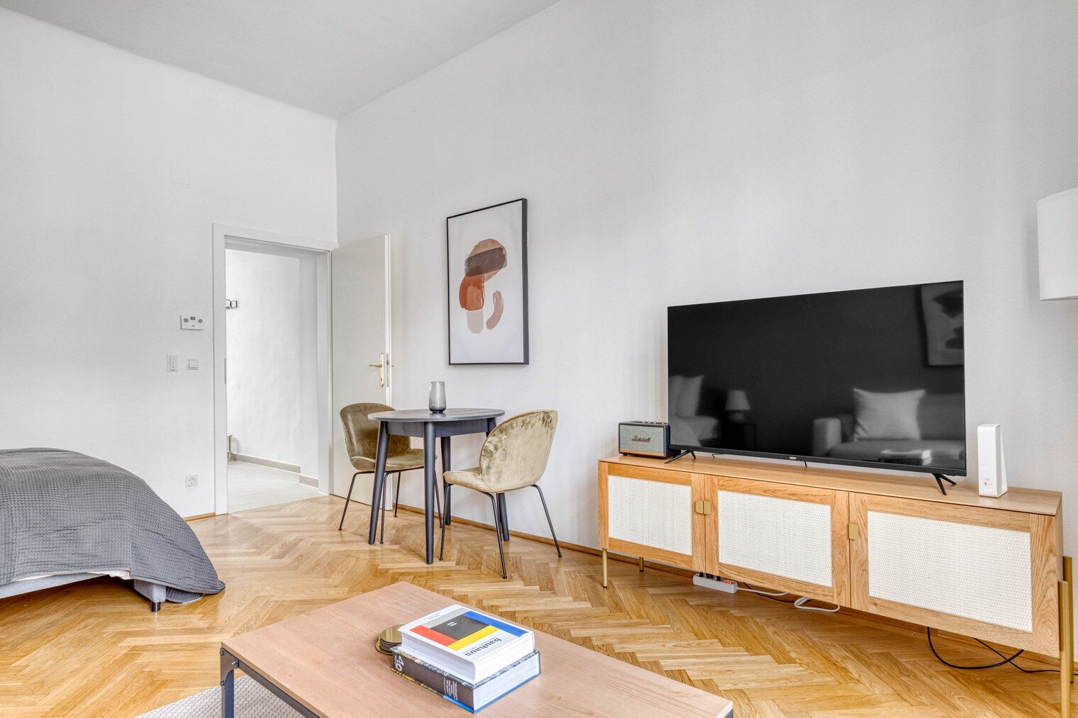Un soggiorno moderno con una grande televisione a schermo piatto, un mobiletto in legno con un altoparlante, un tavolo da pranzo con sedie e un letto con coperta grigia.