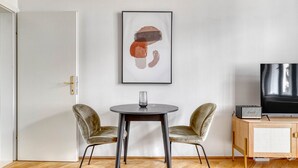 Una habitación con piso de madera en patrón espiga, una mesa redonda con dos sillas de terciopelo, un cuadro abstracto enmarco, un televisor sobre un mueble de madera y una puerta con pomo dorado.