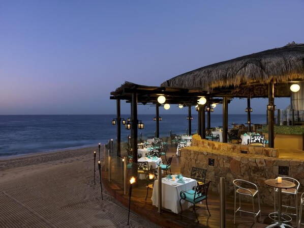 An outdoor dining area with tables and chairs, a thatched roof structure, and a view of the ocean.
