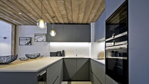 A modern kitchen with a wooden ceiling, grey cabinets, and a white countertop. There are two hanging pendant lights, a sink, and a built-in oven. - Magnificent 8-person Meribel Centre apartment with Nordic bath on terrace (Les Allues)