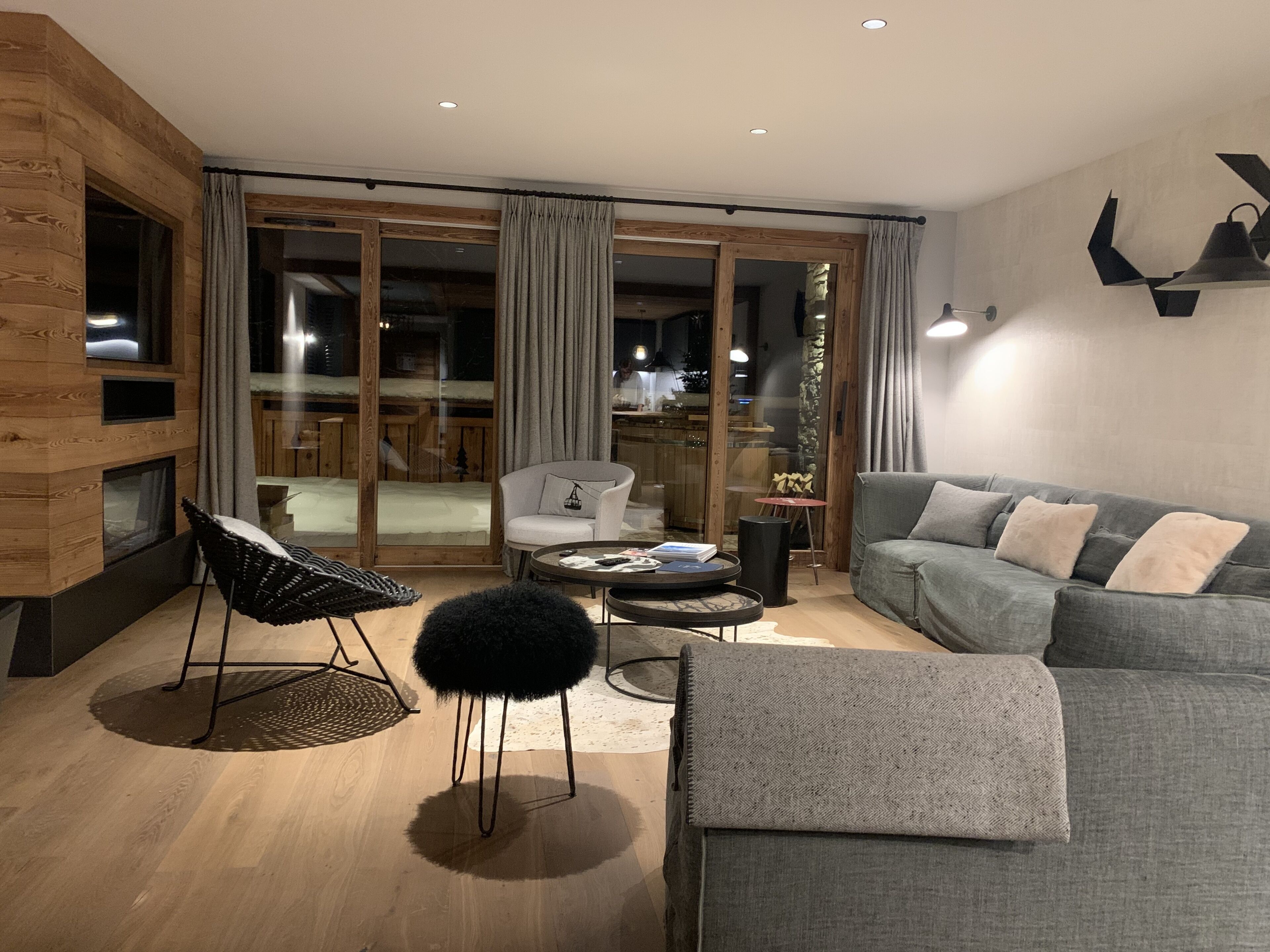 A modern living room with a grey sofa, a white armchair, and a black chair. There is a fireplace, a glass door leading outside, and a dining area visible through the glass.
