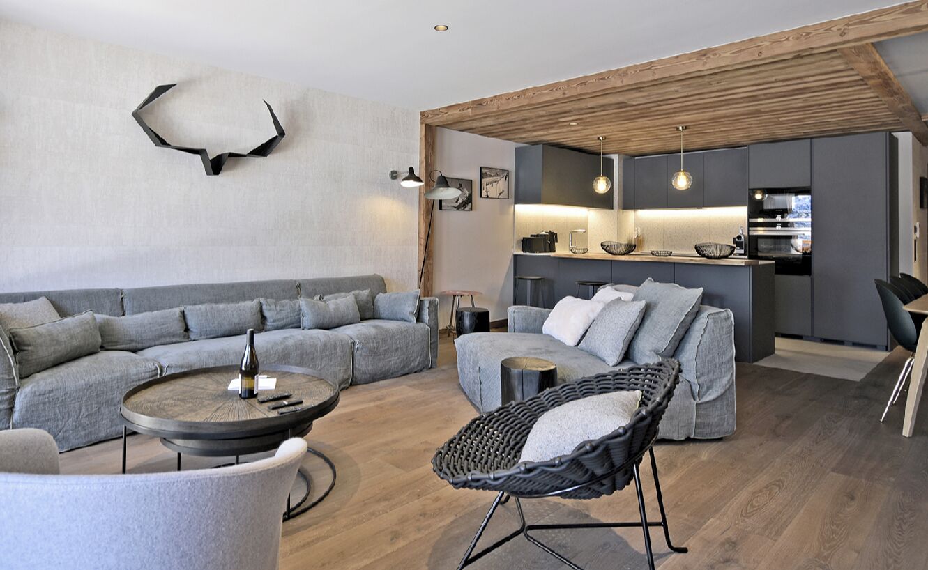 Una sala moderna con un sofá seccional gris, una mesa de centro redonda y una silla de mimbre. Se ve una cocina en el fondo con gabinetes grises y un techo de madera.