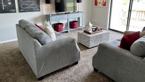 A living room with grey sofas, a coffee table, a TV on a stand, and a poster on the wall. - 28 North Extended Stay Suites!! pickleball, hot tubs! (Traverse City)