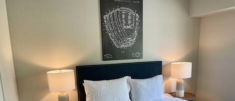 A neatly made bed with white linens, two bedside lamps, a small table with a plant, and a wall-mounted baseball glove diagram.