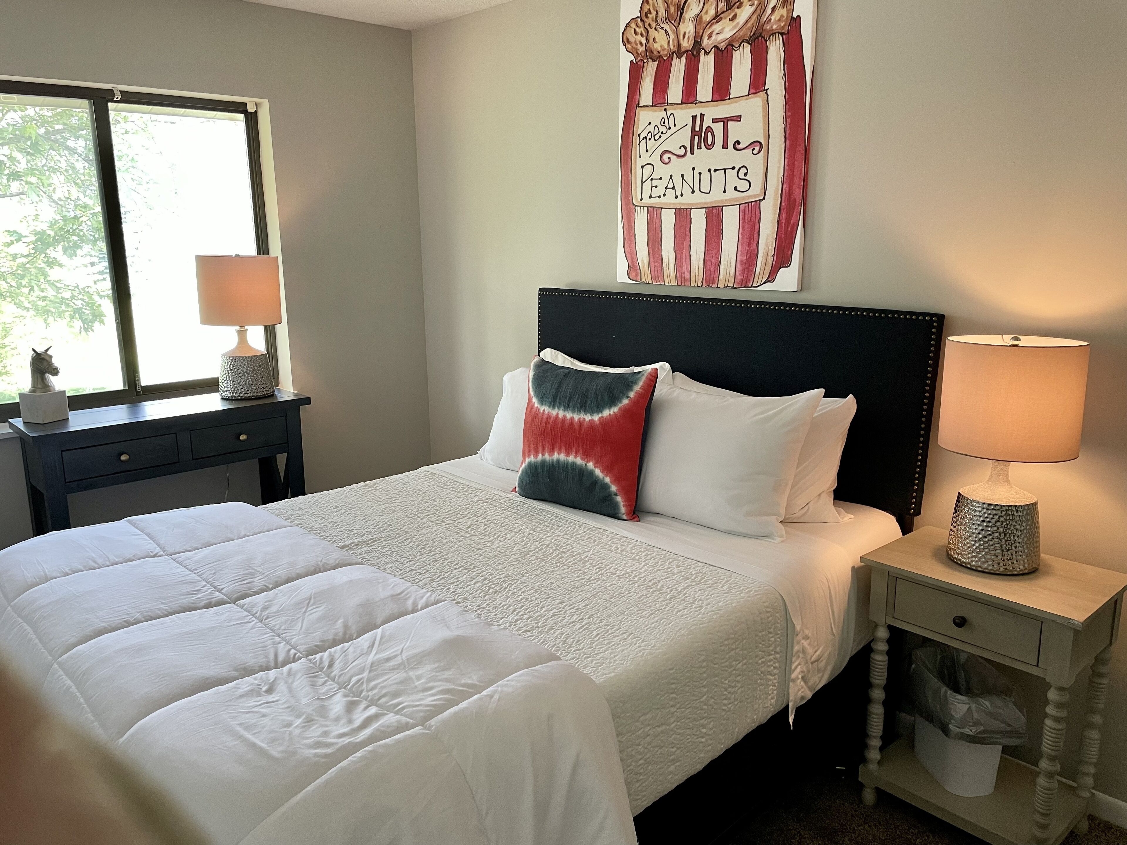 A neatly made bed with white bedding, a headboard with a sign saying "Fresh Hot Peanuts," and a window with a view of greenery.