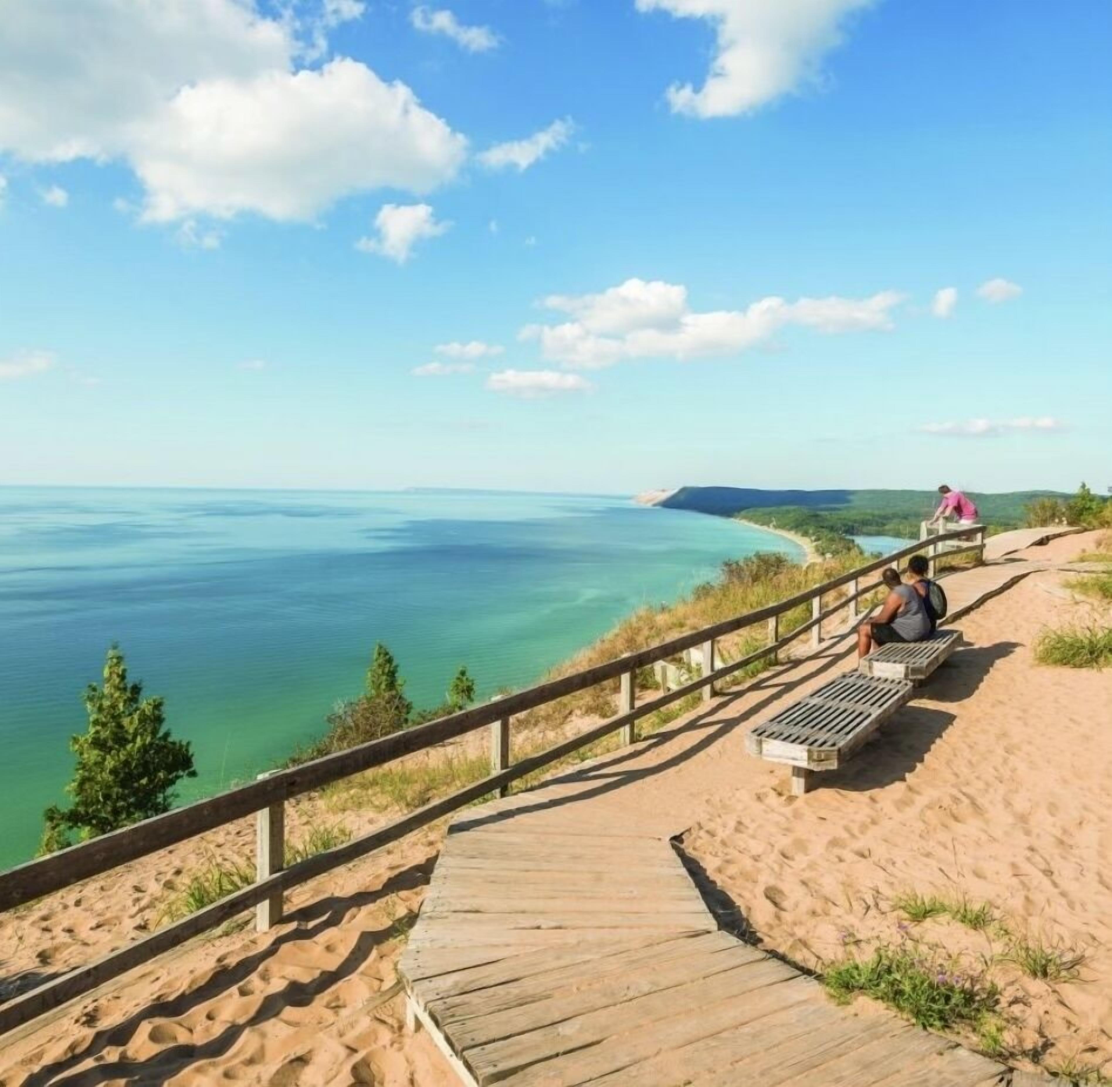 A wooden boardwalk leading to a sandy area with benches, overlooking a clear blue body of water and distant land.