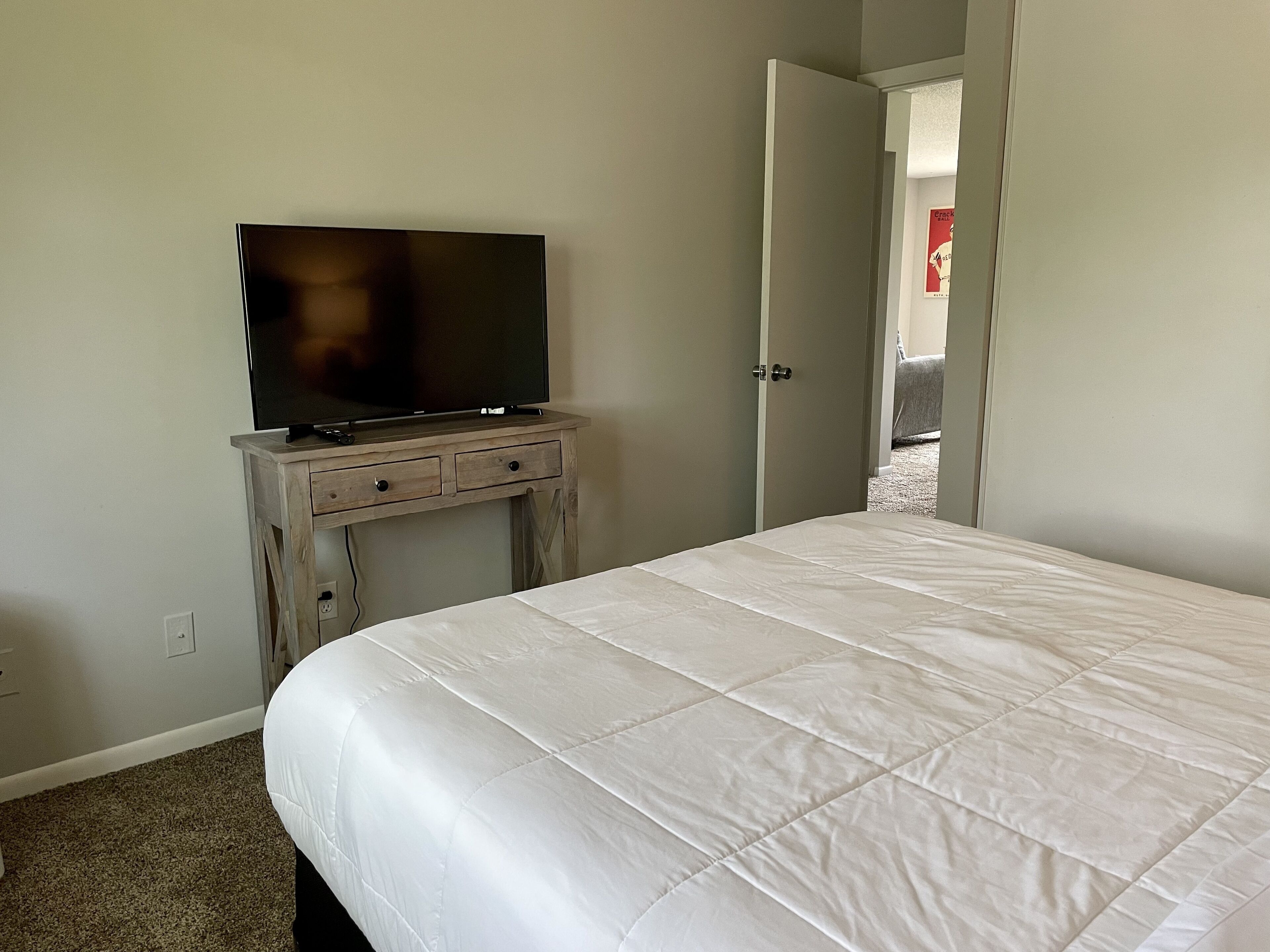 Un dormitorio con cama, televisor sobre un mueble de madera y una puerta que da a otra habitación.
