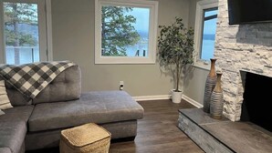 A living room with a grey sectional sofa, a fireplace, a large window with a view of trees and a body of water, and a flat-screen TV mounted on a stone wall. - Modern Lakefront Views at Lake Hickory Haven (Hickory)