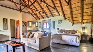 A spacious room with a high ceiling and a thatched roof. There is a sofa and a bed with pillows, a small table, and a lamp.