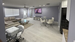 Una sala moderna con una mesa de centro de vidrio, un sofá, una mesa de comedor con sillas, un televisor montado en la pared y una cocina con refrigerador.
