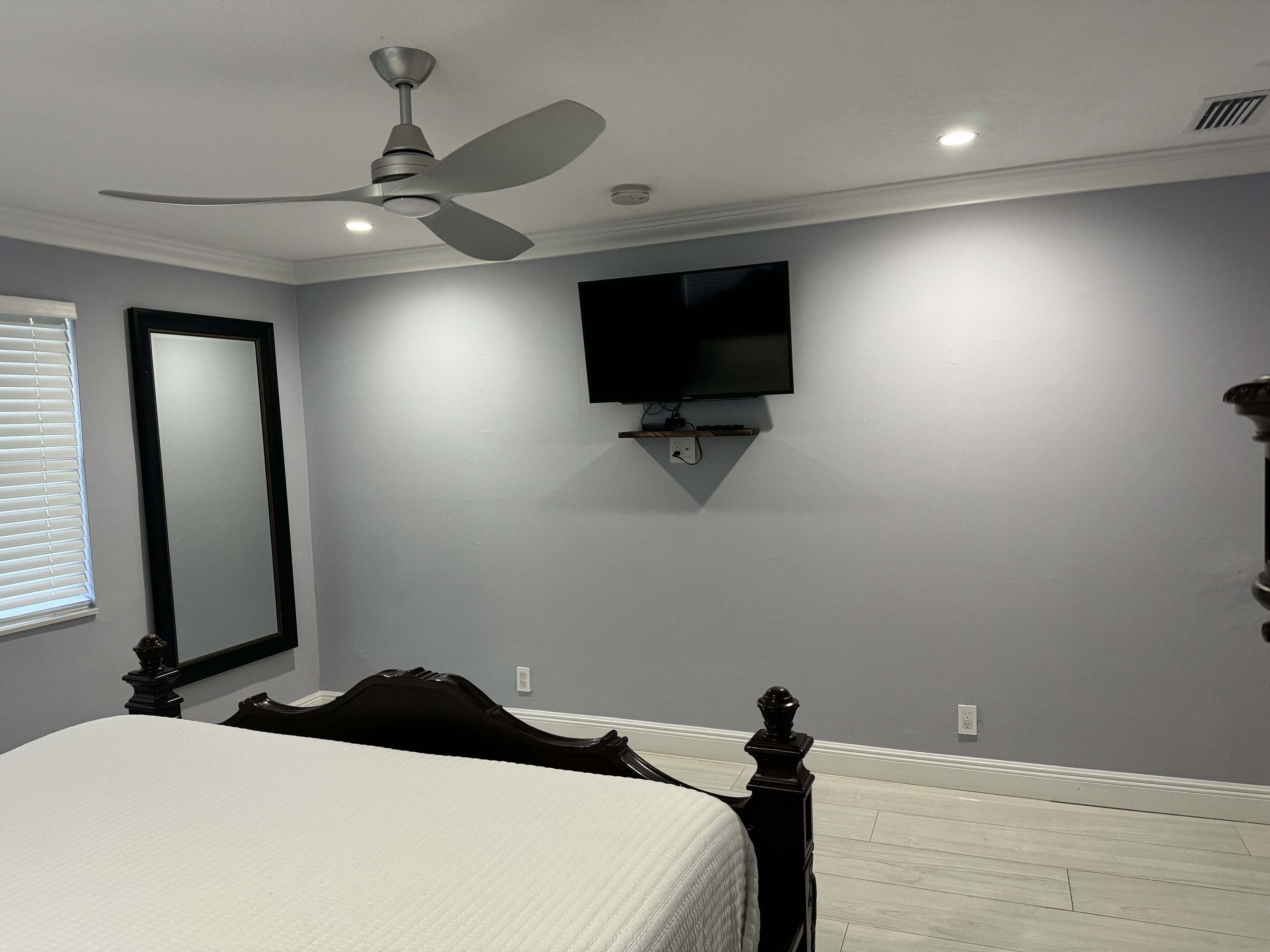 A bedroom with a bed, a ceiling fan, a mounted TV, and a window with blinds.