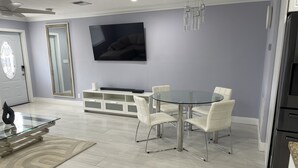 Un comedor moderno con mesa de vidrio y sillas blancas, un banco empotrado con espacio de almacenamiento, y un televisor de pantalla plana grande en la pared.