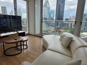 Una sala de estar moderna con un televisor grande, un sofá, una mesita redonda y vistas a la ciudad a través de amplios ventanales.