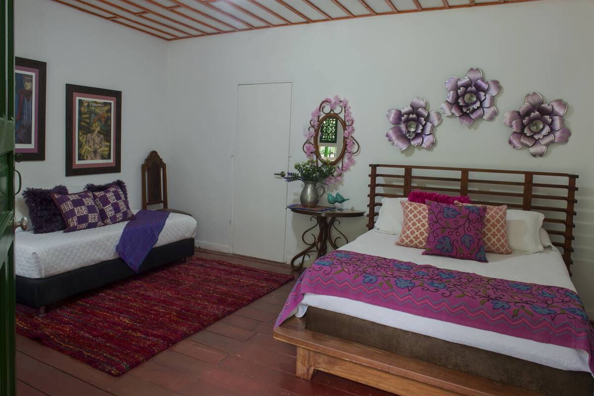 A bedroom with two beds, a wooden headboard, a red rug, and wall decorations including framed pictures and floral wall art.