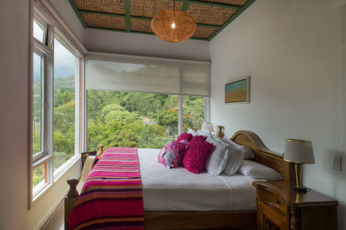 A bedroom with a large window, a bed with white bedding and pink pillows, a wooden headboard, and a bedside table with a lamp.
