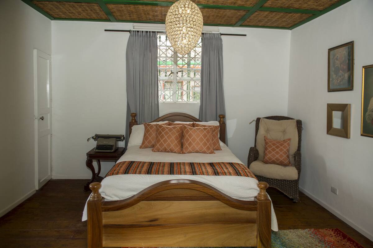 A bedroom with a wooden bed, a chair, a desk with a typewriter, a window with curtains, and a hanging light fixture.