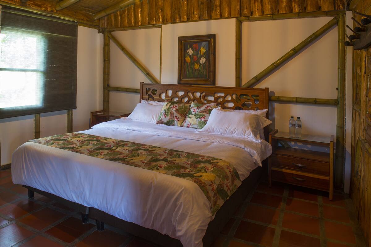 A neatly made bed with floral bedding, a wooden headboard with cut-out designs, a nightstand with a bottle, and a framed picture on the wall.