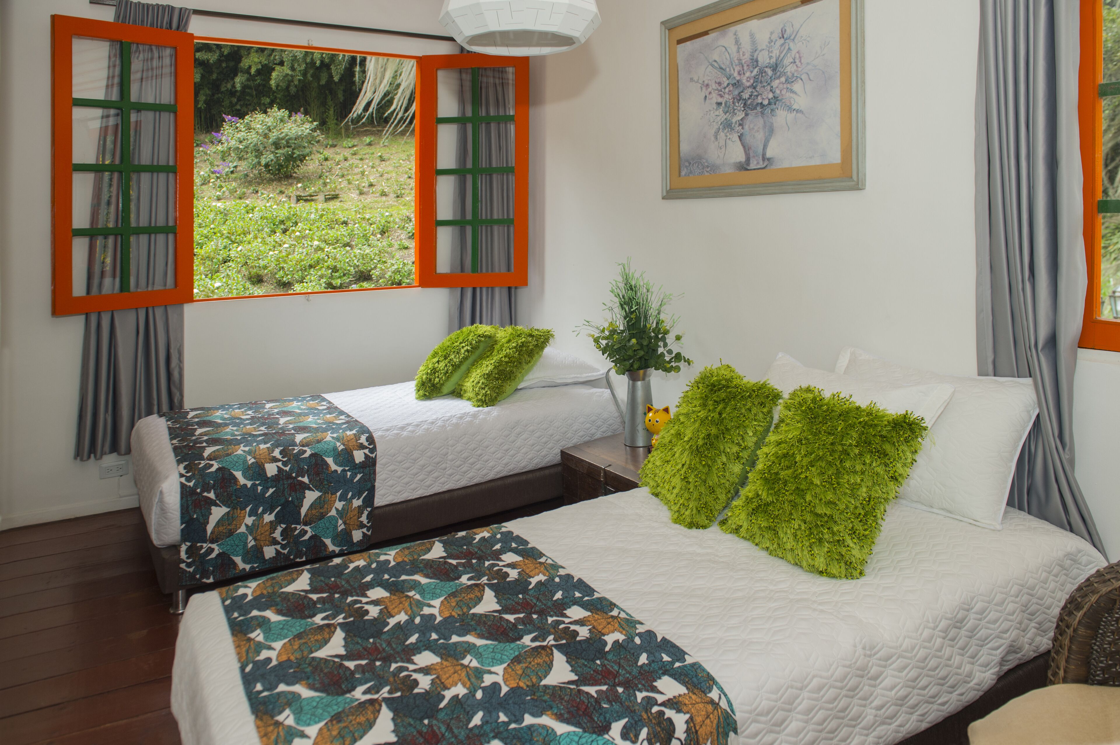 A bedroom with two beds, a window with an orange frame, a painting on the wall, and a small potted plant.