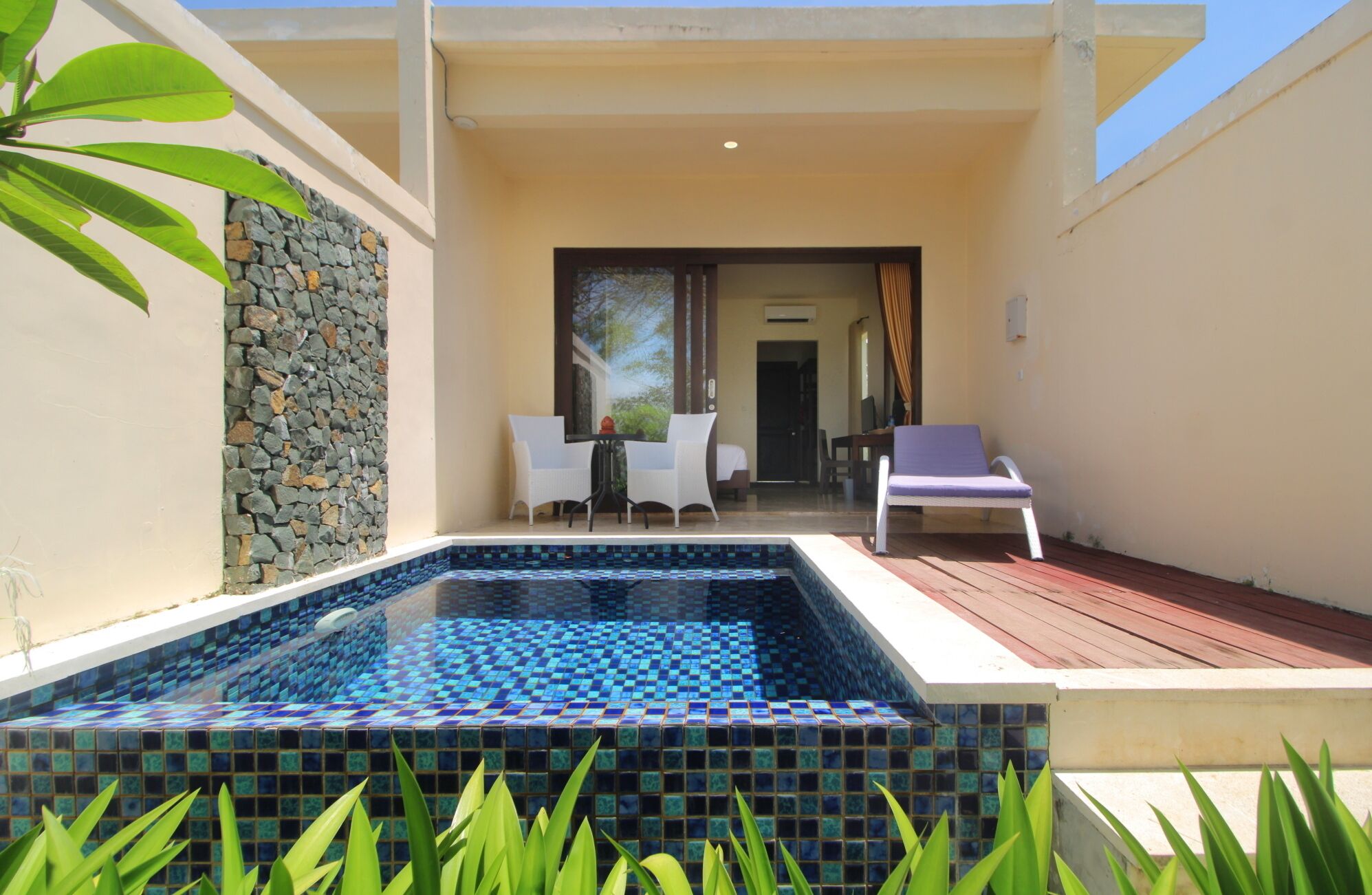 A pool area with a tiled floor, a wooden deck, and a stone wall. There are two white chairs and a lounge chair.