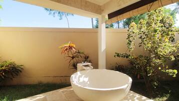 A large white bowl-shaped sink in a sunlit outdoor area with a tiled floor, surrounded by various potted plants and a tree.