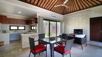 Una cocina moderna con una isla grande, un comedor con mesa de vidrio y sillas de mimbre, y una sala de estar con televisor y sofá.