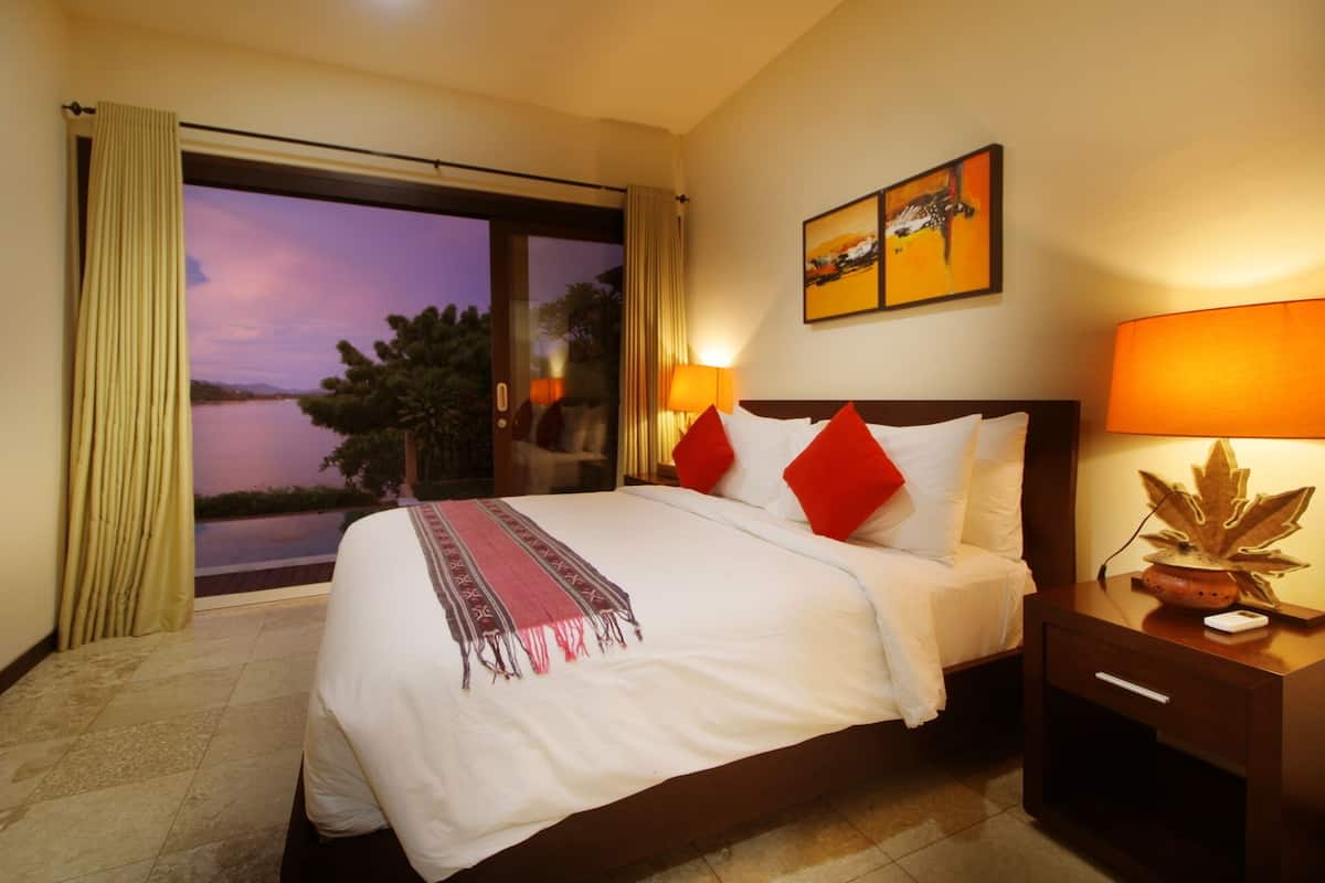 Habitación de hotel con una cama grande, dos almohadas rojas, una mesita de noche con lámpara y vistas al exterior.