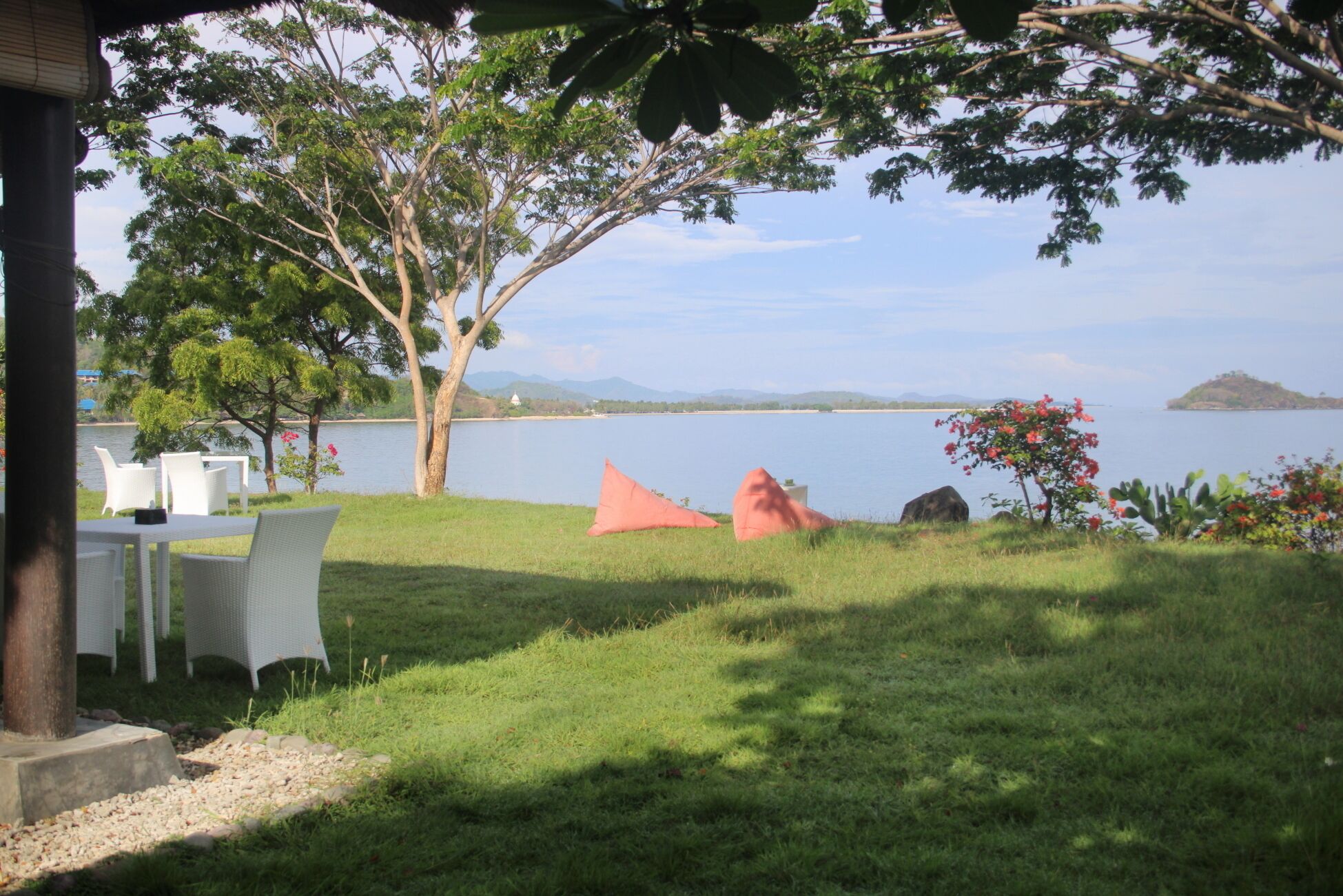 A grassy outdoor area with a table and chairs, a large tree, and a view of a lake with mountains in the background.