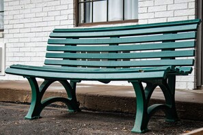 Un banc vert, doté d’un dossier et de deux accoudoirs incurvés, est placé sur un trottoir, devant un immeuble en briques blanches.