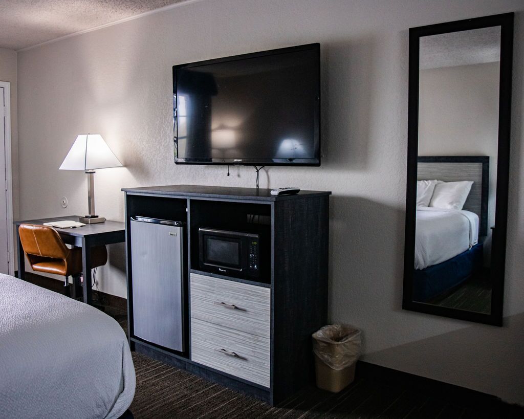 A hotel room with a bed, a desk with a lamp, a minibar, a television, and a mirror.