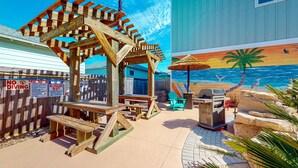 Une terrasse au bord de la plage, agrémentée de structures en bois, d’un barbecue et d’une fresque représentant un paysage de plage.